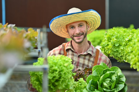Happy male organic farmer holding a wooden crate with fresh organic vegetables sitting in greenhouse plantation.の写真素材