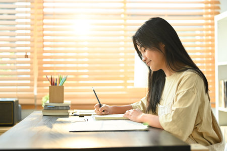 Smiling asian teen girl making notes, ding essay homework assignment at home. Learning and education concept.の写真素材