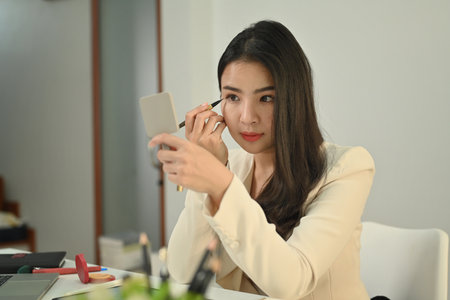 Attractive young businesswoman in stylish suit is making up on working place, applying eyeliner on...
