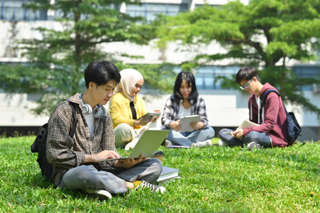 Asian male student using laptop on green grass in campus with friends sitting on background. Education and lifestyle concept.の写真素材