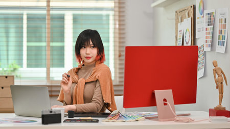 Portrait of young asian woman designer sitting at graphic studio in front of computer monitor and looking confidently to camera.の写真素材