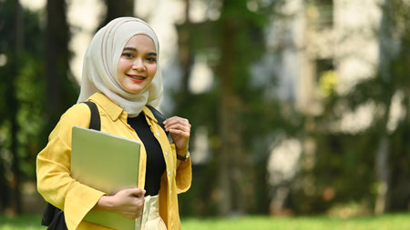 Smiling muslim college student holding laptop standing in the campus. Education, technology and lifestyle concept.の写真素材
