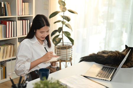 Millennial woman small business entrepreneur writing records to daily planner and using laptop at home office.の写真素材