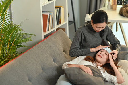 Shot of caring Asian woman taking care of sick girlfriend with fever on couch at home. LGBT, equal...