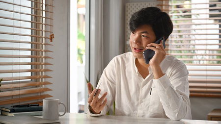 Confident asian male financial advisor consulting client distantly by mobile phone call, sitting at modern workplace.の写真素材