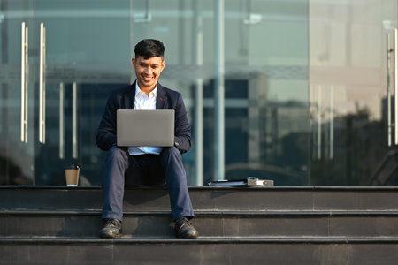 Attractive young businessman sitting on stairs and checking email on laptop before going to the office.の写真素材