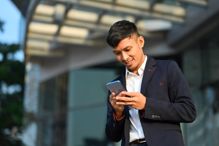 Portrait of handsome man in business suit using mobile phone outside of office in urban city on sunny day.の写真素材
