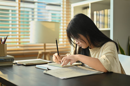 Focused teenaged girl doing homework, writing in notebook while sitting at table in living room.の写真素材