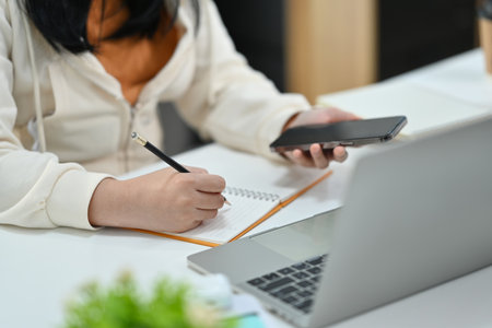 Cropped shot of young woman hand holding mobile phone and making notes on notebook.