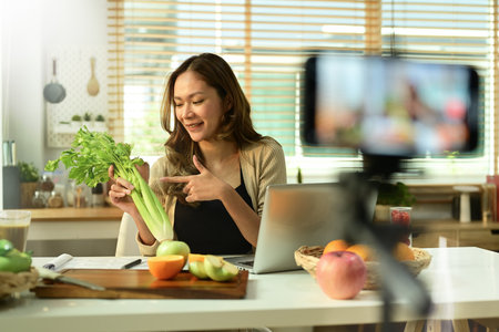 Female nutritionist blogger holding celery, providing with online consultations or recording video content on smartphone.の写真素材