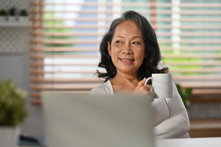 Image of relaxed senior woman in casual clothes enjoys morning coffee and looking through window.の写真素材