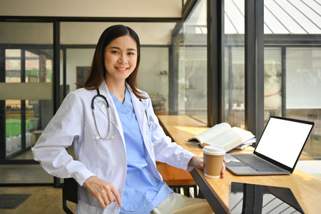 Confident asian female doctor in white coat and stethoscope using laptop in modern hospital canteen.の写真素材