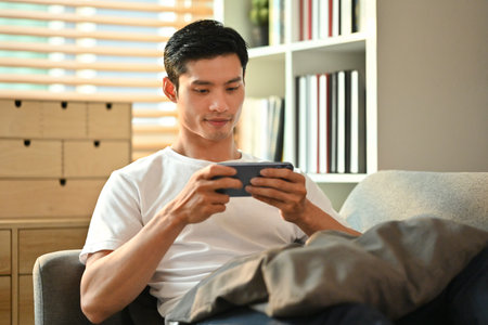 Happy millennial man playing video games on smart phone while resting on couch.Technology, entertainment and leisure activity.の写真素材
