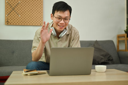 Handsome Asian man making video call on laptop sitting on couch at home. Technology, communicating and lifestyle.の写真素材