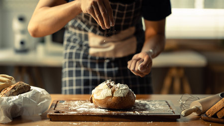 Male baker wearing apron sifting flour on dough, making bread in kitchen.の写真素材