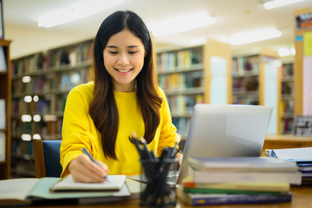 Beautiful Asian college woman preparing report, doing assignments on laptop in a library.の写真素材