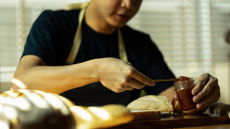 Man preparing a healthy breakfast, spreading raspberry jam on slice of bread with table knife at kitchen table.の写真素材