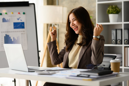 Surprised asian female entrepreneur looking at laptop screen, making winner yes gesture, celebrating business success.の写真素材