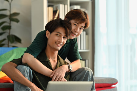 Happy same sex male couple embracing and using laptop in living room. LGBT, love and lifestyle relationship concept.の写真素材