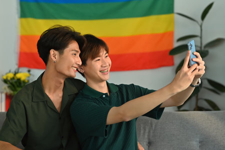 Joyful homosexual couple taking a selfie with smartphone in living room with rainbow flag in background. LGBT and love concept.の写真素材