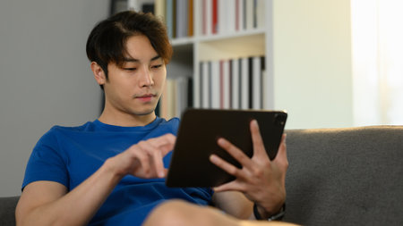 Carefree asian male freelancer sitting on sofa and checking email or working online on digital...
