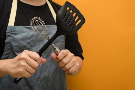 Man wearing apron holding whisks and kitchen spatula standing against yellow background with space.の写真素材
