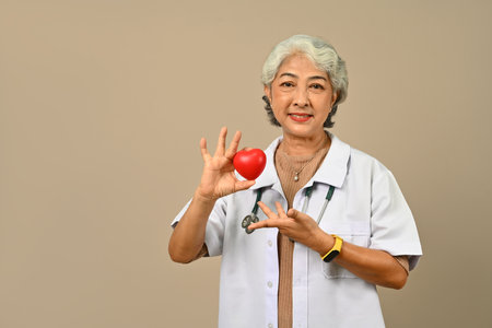 Positive senior woman doctor holding a red heart, isolated on beige background. Cardiology, medical and healthcare concept.の写真素材