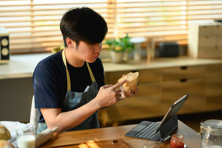 Young asian man wearing casual clothes and apron holding fresh bread and having video call over laptop.の写真素材
