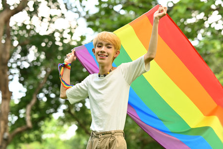 Cheerful young stylish man holding a gay pride rainbow flag blowing in the wind in the park. LGBTQ community, equal rights.の写真素材