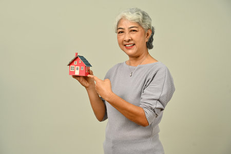 Beautiful mature woman hold small house model isolated on beige background. Real estate, building, property and insurance concept.の写真素材
