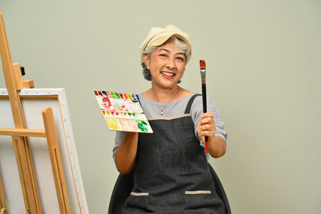 Smiling gray haired senior woman holding palette and paintbrush sitting in front of easel on beige background. People, leisure and hobby.の写真素材