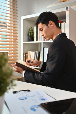 Side view of millennial male entrepreneur reading notes in planner or checking his working schedule plan at workplace.の写真素材