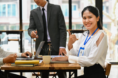 Smiling asian woman employee showing thumb up while working on laptop and her colleagues standing in...