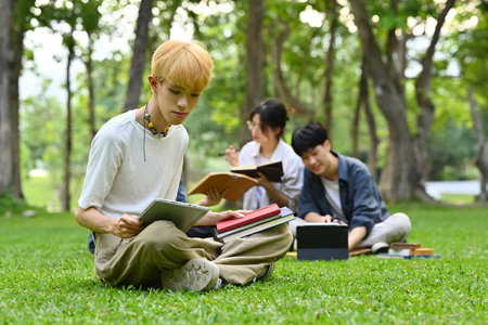 Teenage student man using digital tablet on green grass with friends sitting on background. Education, technology and lifestyle.の写真素材