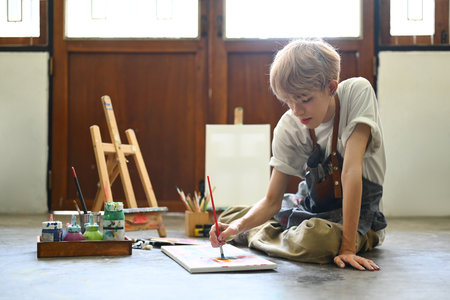 Calm young stylish man sitting on floor in bright art studio and painting picture with oil paints....