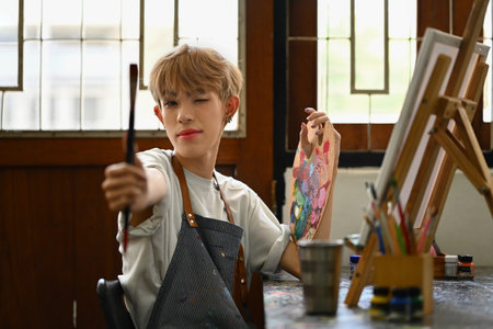 Pretty teenage gay man holding paintbrushes and palette sitting in art studio. Education, hobby, art concept.の写真素材