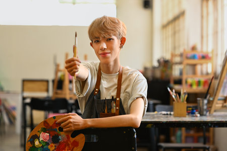 Pretty teenage gay man holding paintbrushes and palette sitting in art studio. Education, hobby, art concept.の写真素材