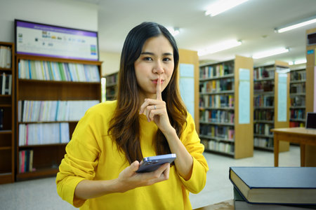 Young woman holding smartphone and making shush gesture as sign of silence, secrecy, secret, keep...