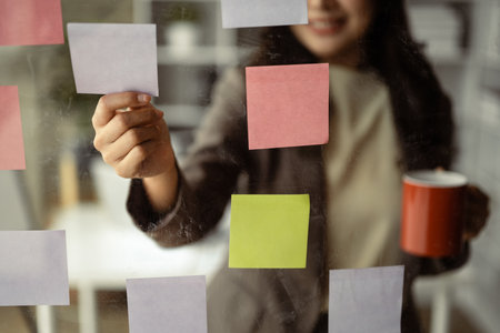 Smiling businesswoman executive manager reading sticky notes on transparent board, planning goals, manage corporate work tasks.の写真素材