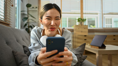 Cheerful young woman lying on sofa in living room using smartphone, texting messaging, communication...