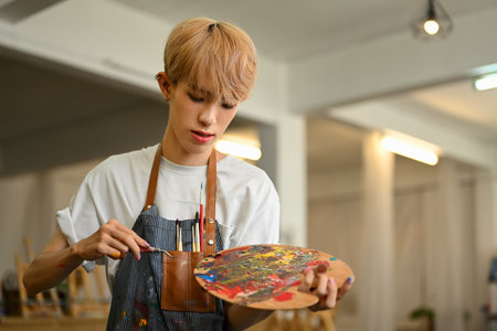 Image of talented gay male artist mixing color oil painting on palette, creating artwork in modern art studio.の写真素材