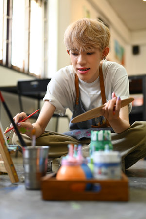 Image of young male artist holding palette and painting picture on canvas with oil paints in art studio.の写真素材