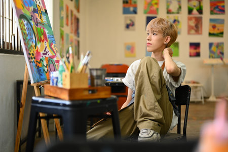 Thoughtful young gay man artist sitting in art studio and looking away, thinking of new creative ideas. Art and education concept.の写真素材
