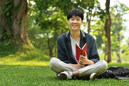 Cheerful asian student man sitting on the grass in the city park and reading a book. Youth lifestyle and education.の写真素材