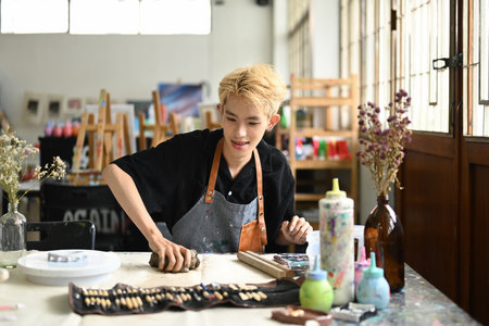 Young creative gay male in apron kneading piece of clay on table in workshop. Handicraft,...