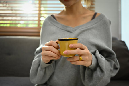 Cropped shot of young woman in casual outfit sitting on couch and drinking herbal tea or coffee.の写真素材