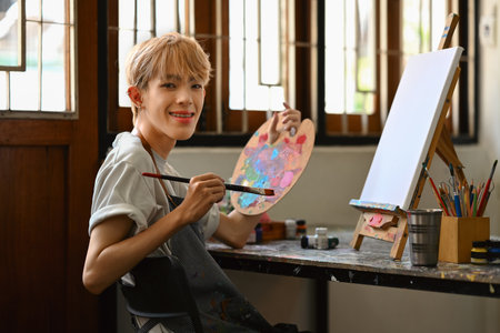 Young hipster queer artist holding palette and brush sitting in front of easel in bright studio....