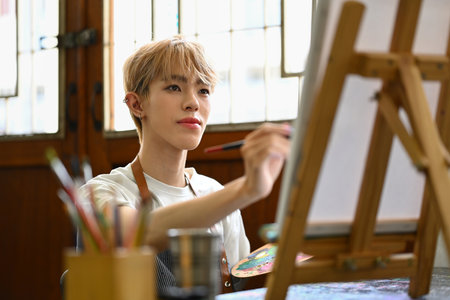 Pleased young gay man painting with watercolor on canvas in art workshop. Art, creative hobby and...