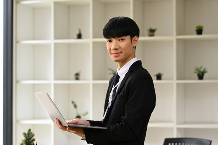 Portrait of young businessman in black suit holding laptop and looking confidently to camera.の写真素材