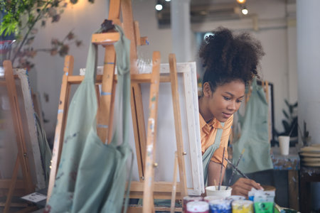 Young African American female artist mixing color oil painting for artwork, sitting in front of easel in art studio.の写真素材